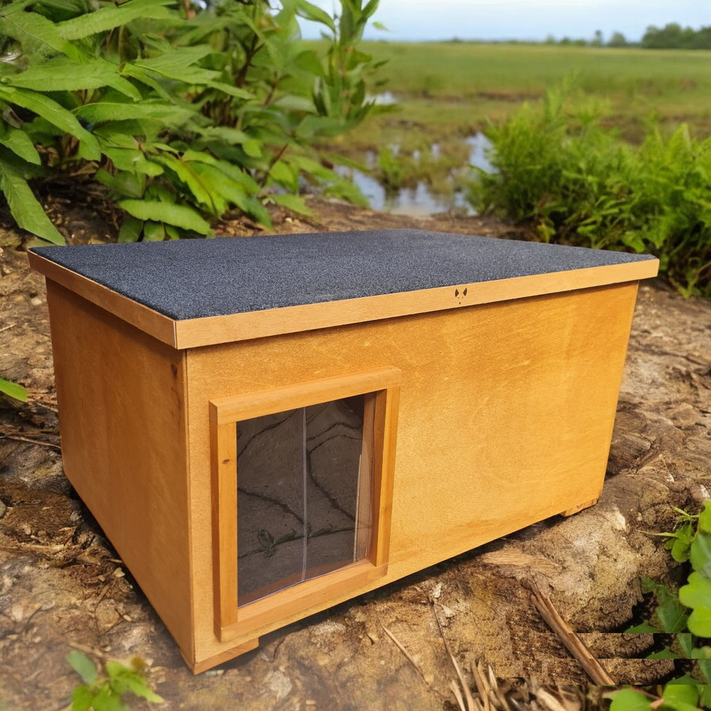 A wooden cat house with a dark gray thermal barrier flap, placed outdoors among plants and rocks.