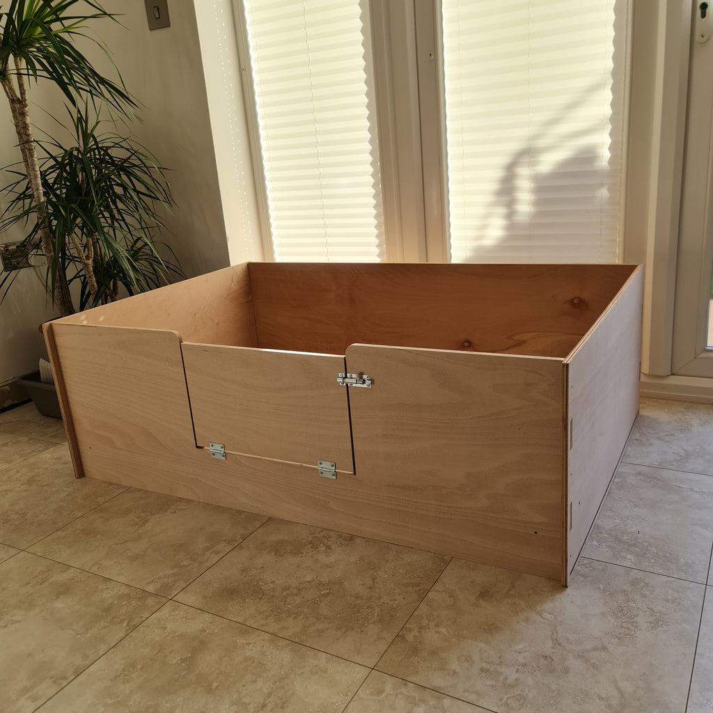 A large wooden whelping box for dogs made from oak plywood, with a large lockable entrance, placed on a tile floor.