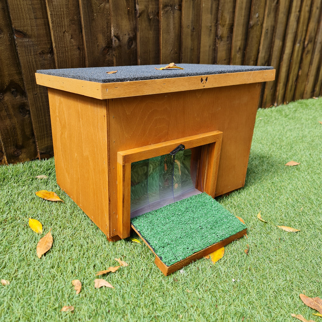 A small wooden insulated tortoise house with a ramp, situated on grass against a wooden fence.