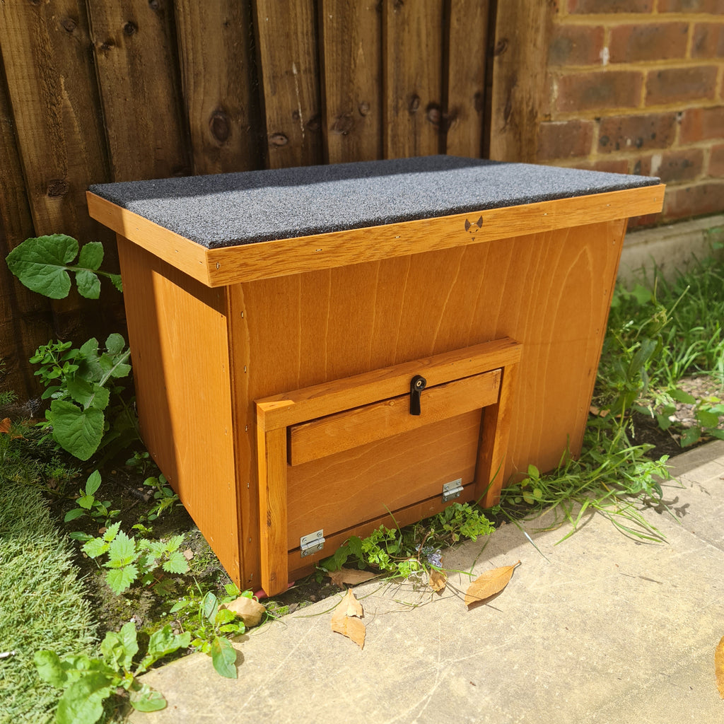 A small wooden tortoise house with a lockable front door and a hinged roof, placed on a garden ground.